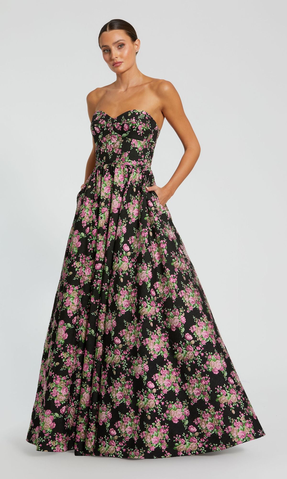 A woman poses against a plain background in the Mac Duggal 68780—a strapless, floor-length black formal dress featuring pink and green floral prints.